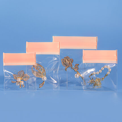 Four pink clear EVA jewelry storage ziplock bags in graduated sizes standing upright, each containing gold brooches and pearl bracelets
