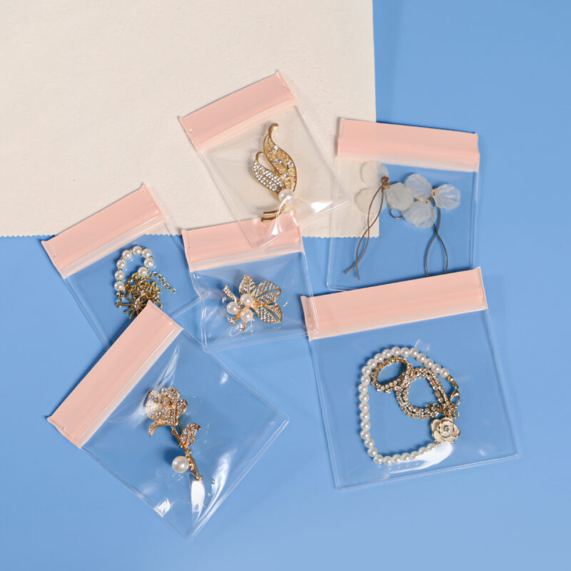 Six transparent EVA jewelry pouches with pink ziplock closures laid flat, holding assorted jewelry including brooches, bracelets and flower accessories, showing size variety