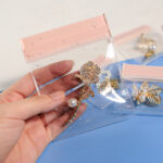 Hand holding a waterproof pink EVA transparent jewelry ziplock bag covered in water droplets, containing a crystal rose brooch inside, with additional jewelry bags in background