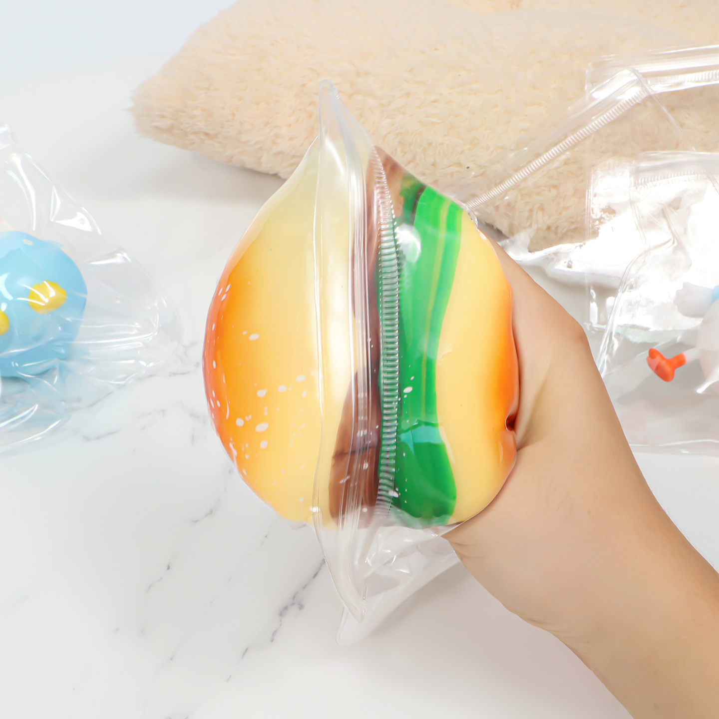 Hand forcefully squeezing a hamburger squishy inside a flexible squishy toy packaging bag to demonstrate film and seal durability.
