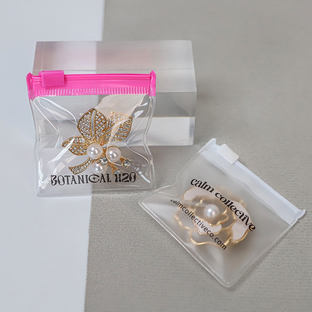 Custom printed clear plastic jewelry bags with colored zippers, displaying brand logos and a pearl brooch inside.